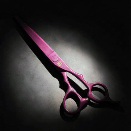 A pair of metallic pink hair shears with a gold logo on the blade, set against a dark background with dramatic spotlighting.