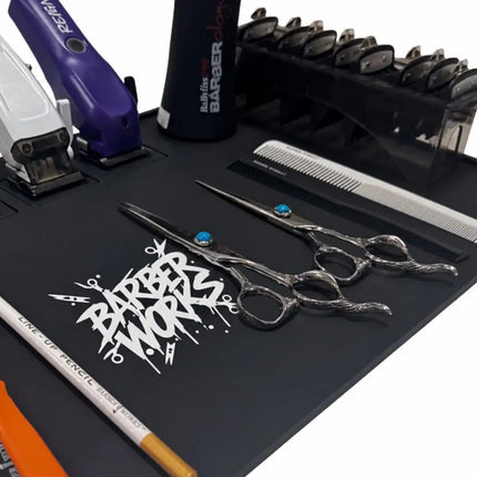 A barber station set on a black mat, featuring silver scissors with blue accents, a white comb, a black clipper, a purple trimmer, and a black cordless clipper holder. The mat has a white 'BARBER WORKS' logo printed on it.