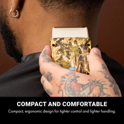 BaBylissPRO Camo LO-PROFX Compact Single-Foil Shaver (Limited Edition) Hand holding a compact electronic device with a tattooed arm, against a brown background.
