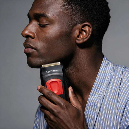 A close-up of a man with closed eyes holding a black and red Gamma+ electric trimmer against his neck, wearing a blue and white striped shirt against a solid gray background.