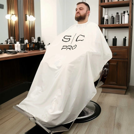 SC Pro White Cape with Black Logo (Kick Capes) Man sitting in a barber chair with a white cape in a salon setting.