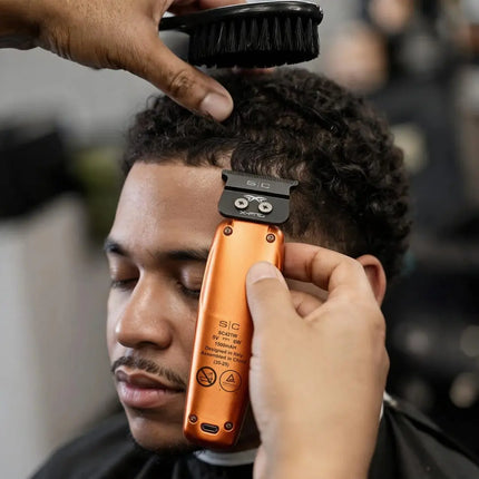 A close-up of a person getting a haircut with a barber using an StyleCraft Saber Professional Hair Trimmer – Limited Edition Orange on curly dark hair. A hand is brushing the hair while the other holds the clipper, with a blurred barbershop background.