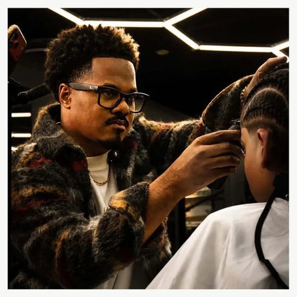 A man wearing a dark patterned faux fur jacket and black-rimmed glasses styles a child's hair in a barbershop setting with geometric ceiling lights.