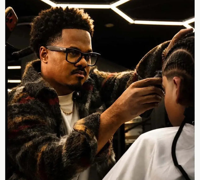 A man wearing a dark patterned faux fur jacket and black-rimmed glasses styles a child's hair in a barbershop setting with geometric ceiling lights.