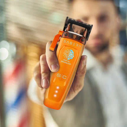 A close-up of a person holding an orange StyleCraft Saber 2 Professional Hair Clipper – Limited Edition Orange against a blurred indoor background.