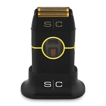 A black and gold Instinct Metal Shaver with a visible brand logo on the front, housed in a charging stand with a clear protective cap on top.