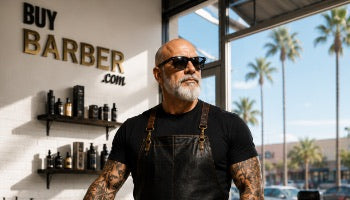 A tattooed man with a beard and sunglasses stands inside a barbershop, wearing black overalls over a black t-shirt. The background features a white wall with 'BUY BARBER .