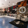 the Barber Company, buybarber.com las vegas store interior with shelves of products, wall-mounted jerseys, an American flag, guitars, helmets, and a large circular sign reading 'The Barber Company.' Two cream armchairs sit on a black rug with a skull-and-triangle logo.