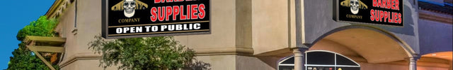 A storefront building for The Barber Supplies, with two large black signs featuring a skull-and-crossbones logo and red and white lettering, set against a blue sky with landscaping and the entrance visible.